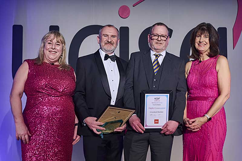 4 people dressed up stood on a stage one person holding an a4 certificate in a fram