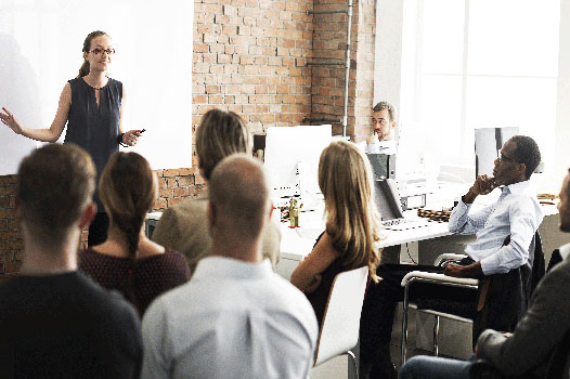 room of adults sitting with a woman stood at front pointing at a white board