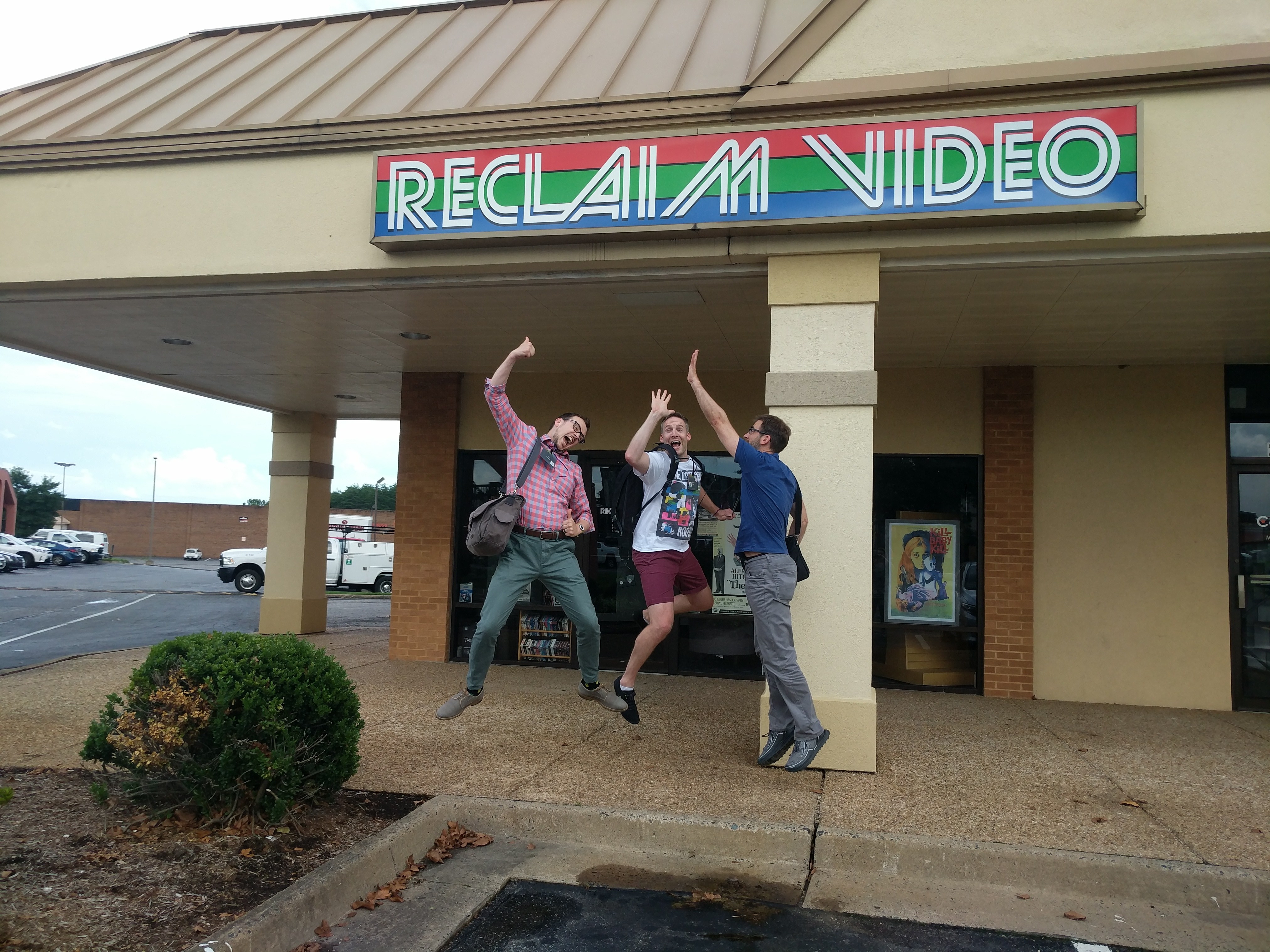 Colour photograph of three men performing a freeze frame high 5 outside an 80s inspired video shop